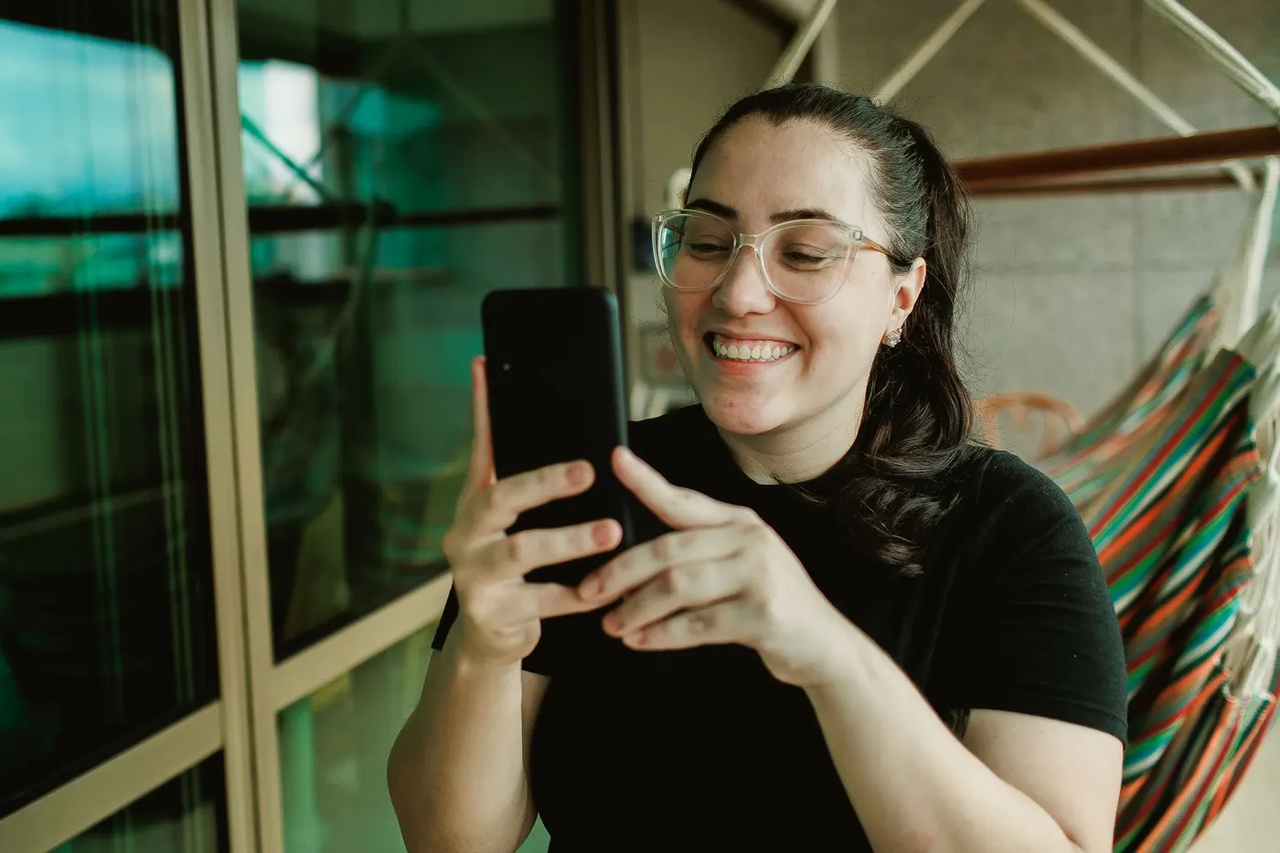 Mulher de óculos, sorrindo ao olhar para a tela do celular, na varanda de casa